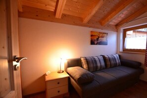 A cozy room with a wooden ceiling, a gray sofa with a plaid pillow, a bedside table with a lamp, and a painting on the wall. - Holiday home in Allgäu with 3 bedrooms (Lechbruck am See)