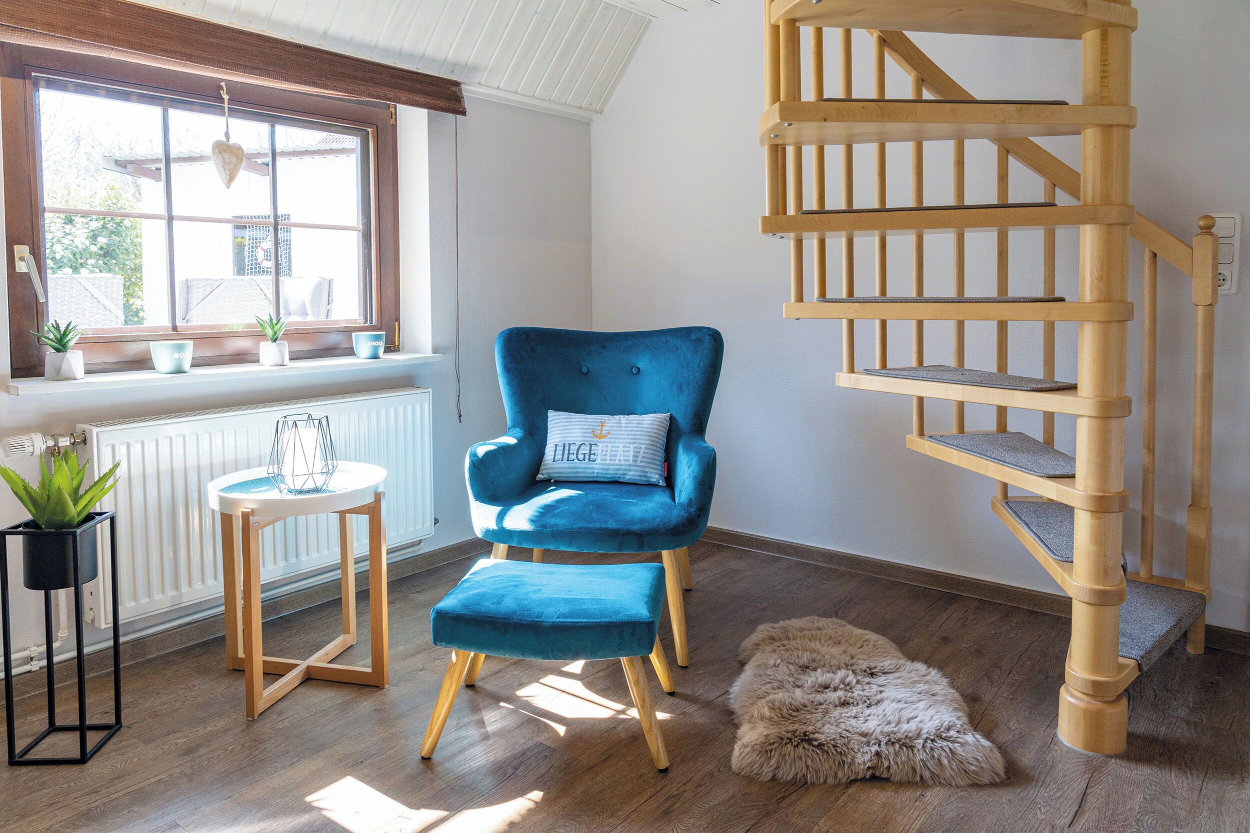 A room with a blue chair, a small stool, a wooden spiral staircase, and a window with plants.