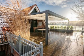 A covered outdoor area with a wooden deck, a brick building, and a view of a body of water. - Country Bliss - Nordhorn (Nordhorn)