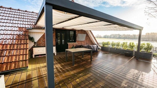 Un espace extérieur abrité, doté d’une table et de chaises, un bâtiment en briques et une vue sur une étendue d’eau.