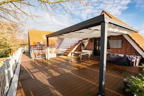 A covered outdoor deck with a table and chairs, a brick wall, and a wooden roof structure. - Country Bliss - Nordhorn (Nordhorn)