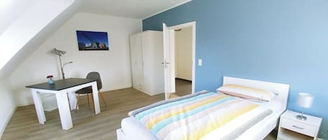 A bedroom with a bed, a desk, and a chair. There is a wardrobe and a door leading to another room.