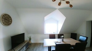 A modern living room with a flat-screen TV, a dining table with chairs, a clock on the wall, and a unique geometric ceiling light fixture. - Holiday apartment Klever Berg (Kleve)