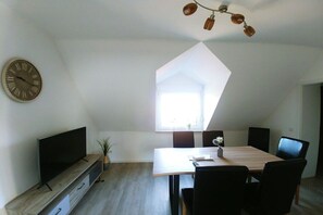 A modern living room with a flat-screen TV, a dining table with chairs, a clock on the wall, and a unique geometric ceiling light fixture. - Holiday apartment Klever Berg (Kleve)