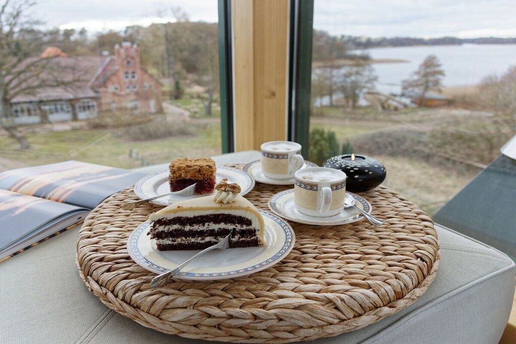 Ein Korbtablett mit einem Kuchenstück und zwei Tassen Tee auf einem Tisch vor einem See mit Blick durch ein Fenster.