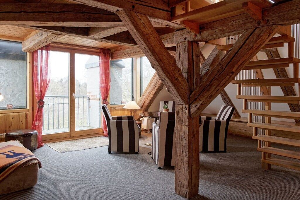 A room with a sloped ceiling, wooden beams, a sofa, a chair, and a window with curtains.