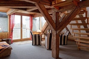 A room with a sloped ceiling, wooden beams, a sofa, a chair, and a window with curtains. - Holiday room with 1 bedroom (Groß Zecher)