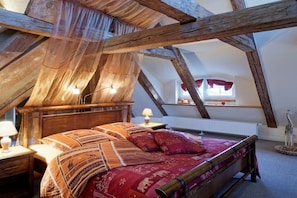 Ein Schlafzimmer mit geneigter Decke, Holzbalken, einem Bett mit gemusterten Textilien, einem Nachttisch mit Lampe und einem Fenster mit Vorhängen.