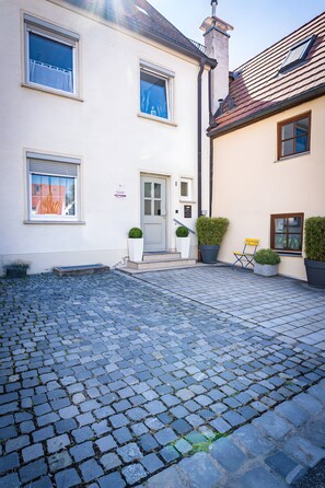 A cobblestone courtyard with a white building featuring a door and windows, potted plants, and a chair.