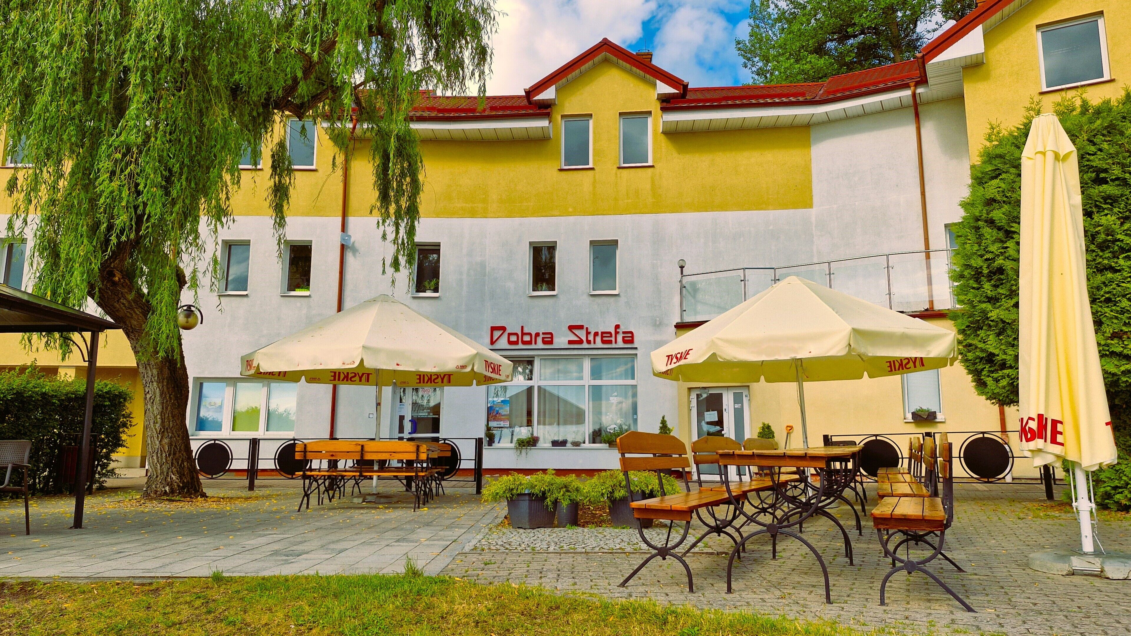 An outdoor seating area with tables and chairs, a building with a yellow facade, and a sign reading 'Dobra Strefa'.