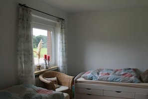A bedroom with a window, a wicker chair, a bed with a patterned blanket, and a bedside table with candles. - Holiday home in Aurich with swimming pool (Aurich)