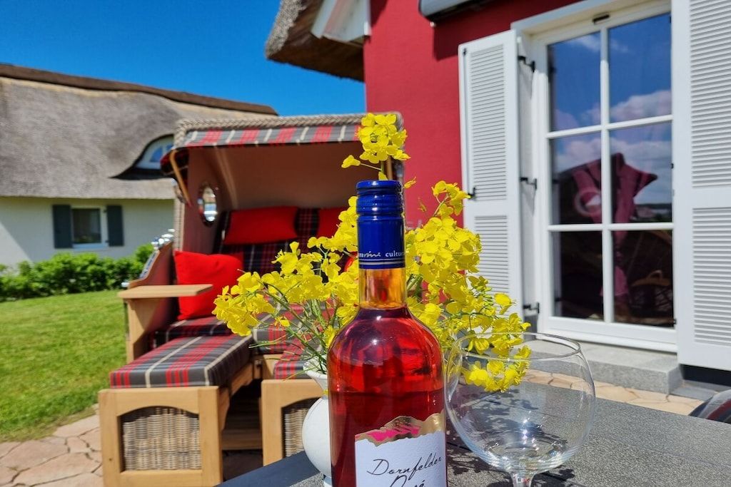 Eine Flasche Dornfelder Wein, ein Glas und gelbe Blumen auf einem Tisch vor einem Gebäude mit roter Fassade und weißen Fensterläden.