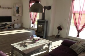 Ein Wohnzimmer mit Fernseher, einem Holztisch mit Blumen, einer Couch, einer Wandheizung und einem Fenster mit Vorhängen.