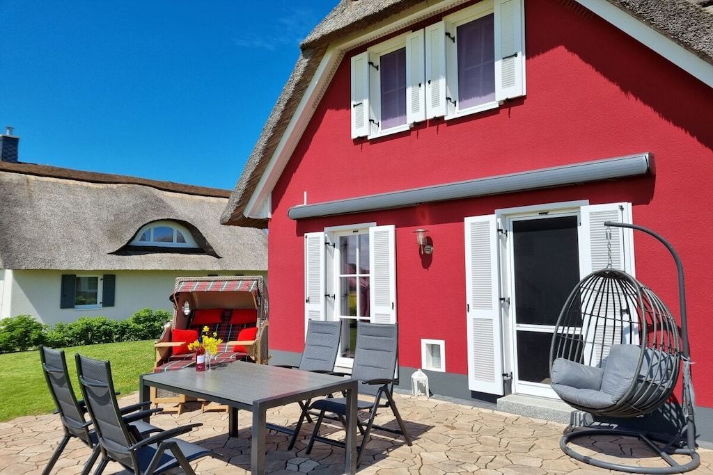 Un bâtiment rouge avec un toit de chaume, une terrasse avec une table et des chaises, et un fauteuil suspendu.