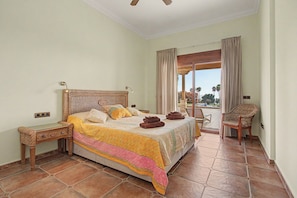 A hotel room with a bed, a chair, a balcony, and a view of palm trees.