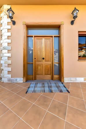 A wooden door with a glass panel, flanked by two wall-mounted lanterns, set against a beige wall with a tiled floor. - Villa Samarin – Golf & Ocean View (Adeje)