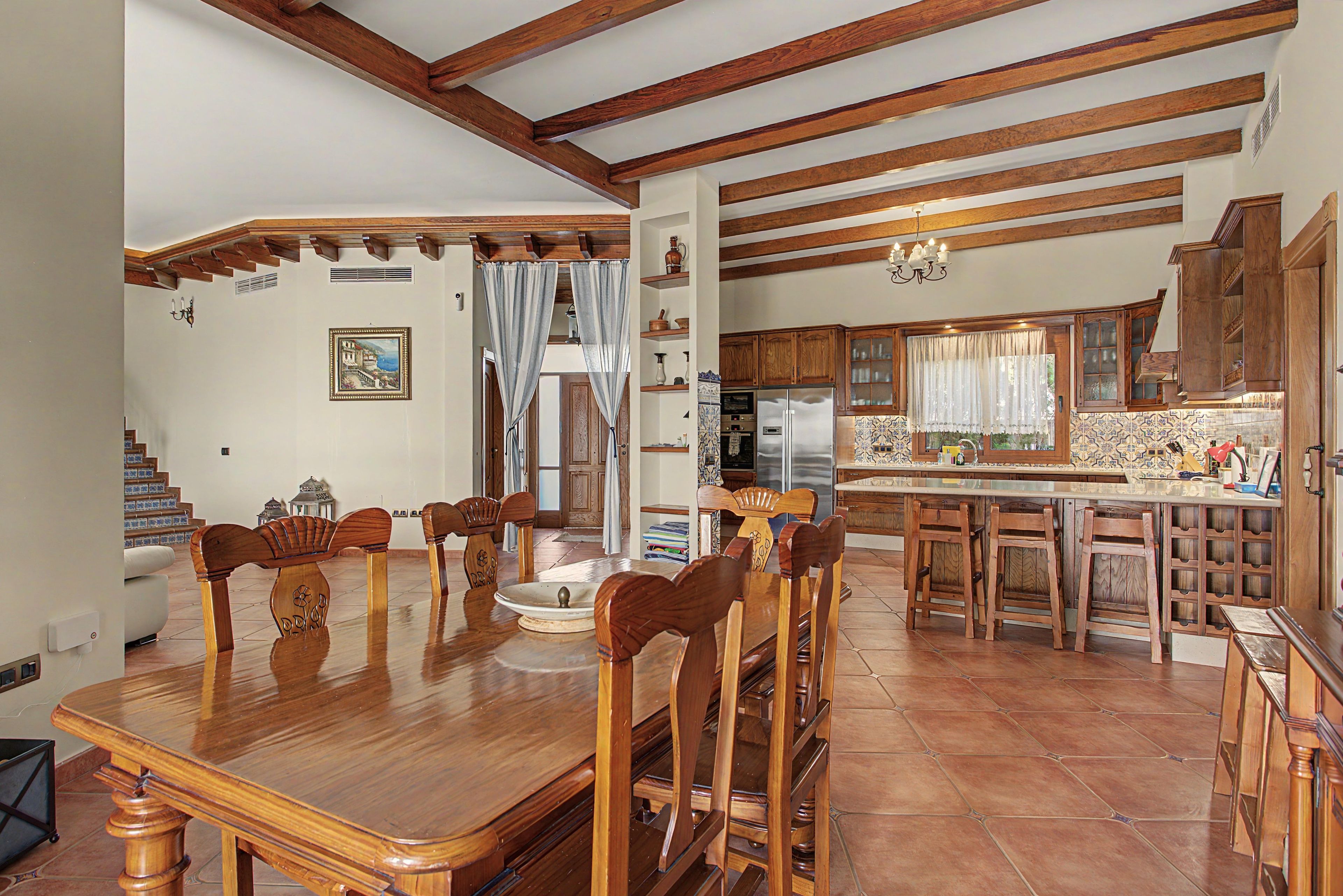 A spacious dining area with a wooden table and chairs, a kitchen with wooden cabinets and a tiled backsplash, and a staircase leading to another level.