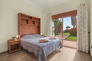 A bedroom with a bed, wooden headboard, nightstands, and a view of a garden through a glass door. - Villa Samarin – Golf & Ocean View (Adeje)