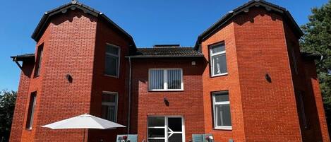 A two-story red brick building with a central entrance, a covered patio area with outdoor seating, and a clear blue sky.