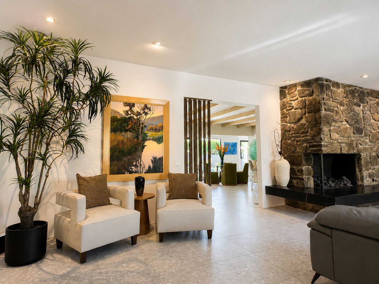 A modern living room with a stone fireplace, two armchairs, a large potted plant, and a painting on the wall.