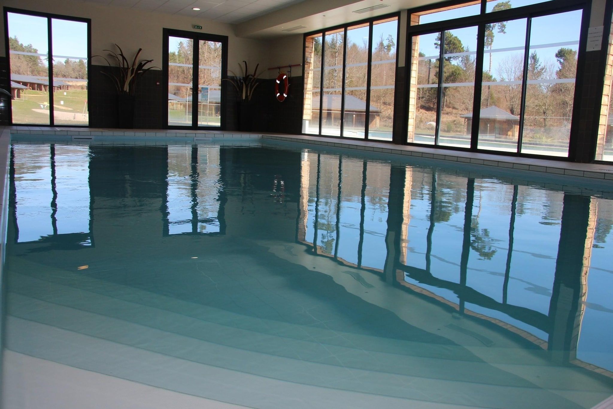 Una piscina interior con paredes de vidrio transparente, piso de baldosas y vistas al exterior a través de amplias ventanas.