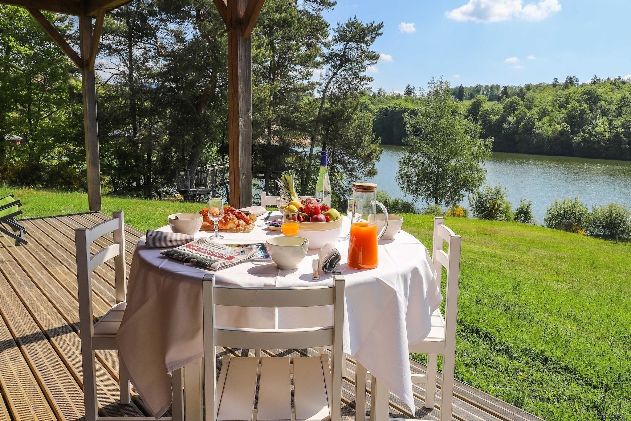 En uteservering med ett bord för två personer, omgiven av ett trädäck, med utsikt över en sjö och omgivande träd och grönska.