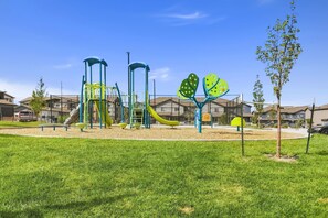 A playground with slides and climbing structures, surrounded by a grassy area and residential buildings.
