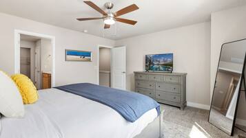Un dormitorio con cama, ventilador de techo, cómoda, espejo y una puerta que da a otra habitación.