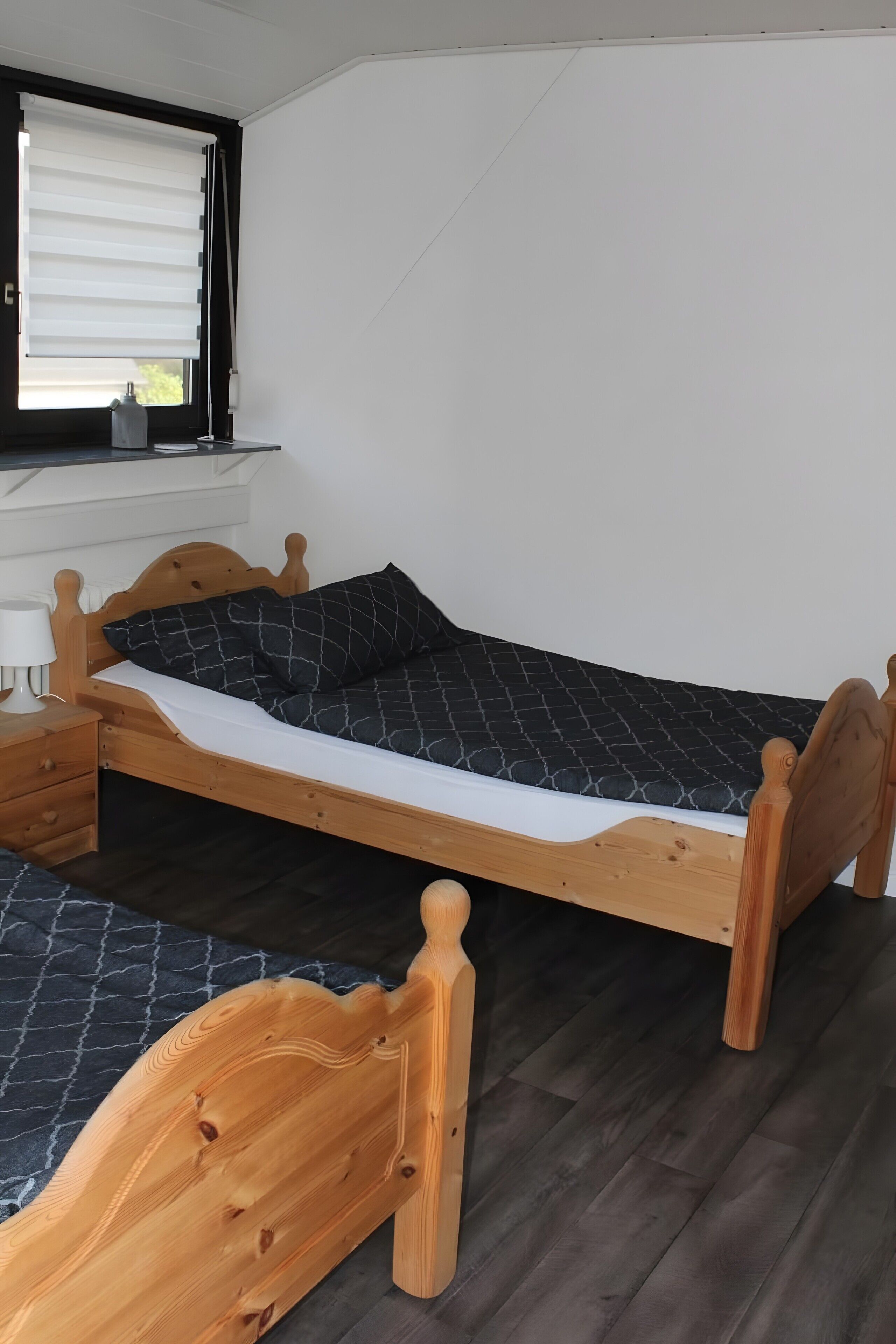 A single wooden bed with a mattress, a bedside table with a lamp, and a window with blinds.
