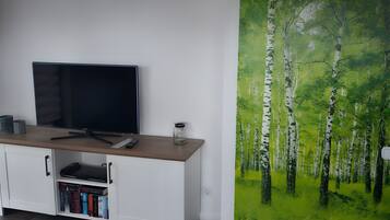 Ein Zimmer mit einem Flachbildfernseher auf einer Holzkommode, einem Bücherregal mit Büchern und einem großformatigen Wandbild eines Waldes.