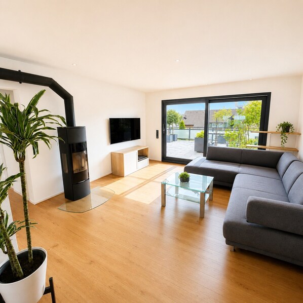 A modern living room with a sofa, a glass coffee table, a flat-screen TV, a fireplace, and a view through large sliding doors to an outdoor area. - Apartment 'Haus Biggeglück' with Private Roof Terrace & Walk to Lake Biggesee (Olpe)
