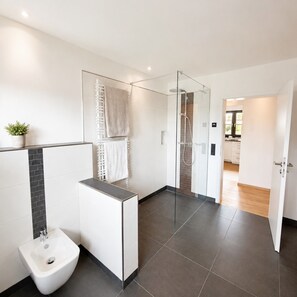 A modern bathroom with a bidet, a glass shower enclosure, and a view into the kitchen. - Apartment 'Haus Biggeglück' with Private Roof Terrace & Walk to Lake Biggesee (Olpe)