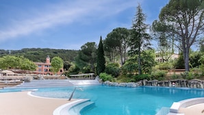 Una piscina amb una font d'aigua cristal·lina i blava, envoltada d'una zona verda amb arbres i un edifici al fons.