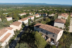 Une vue aérienne d’un quartier résidentiel, avec des maisons aux toits de tuiles rouges, entourées de verdure et surplombant une chaîne de montagnes lointaine.