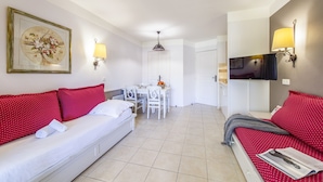 Habitació d'hotel amb un sofà blanc, coixins vermells amb punts, una taula de menjador amb cadirees, un televisor fixat a la paret i un llum al sostre.