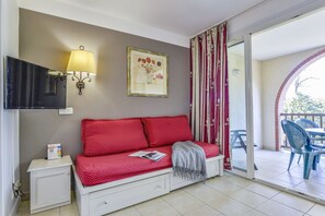 A room with a red sofa, a small bedside table, a wall-mounted lamp, a framed picture, and a sliding glass door leading to an outdoor area with chairs. - Ardèche Holiday Apartment for 4 (Grospierres)