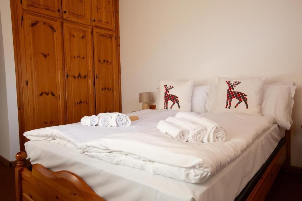 Una cama bien hecha con ropa blanca y toallas enrolladas, un armario de madera y dos almohadas con diseño de reno.