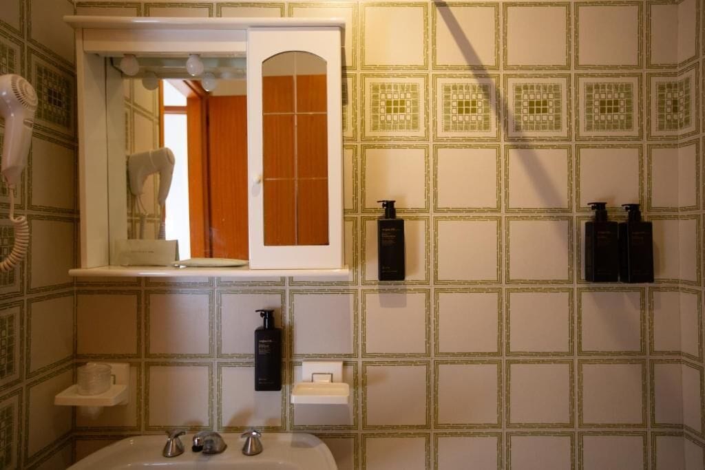 Ein Badezimmer mit Waschbecken, zwei Haartrocknern und einem Spiegel mit geöffneter Tür, hinter dem sich eine Holztür befindet.
