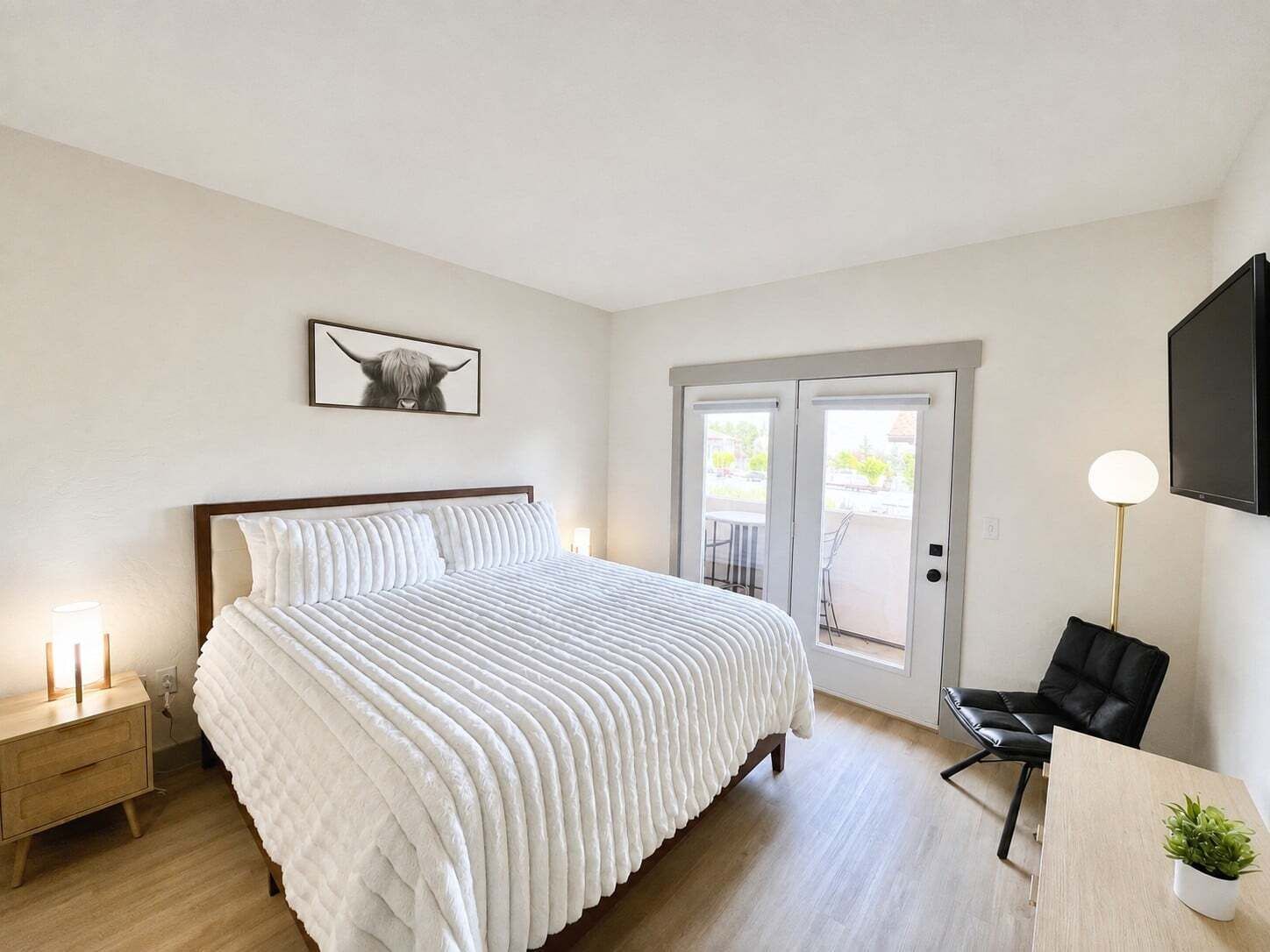 A hotel room with a bed, bedside tables, a desk, a chair, a TV, and a door leading to an outdoor area.