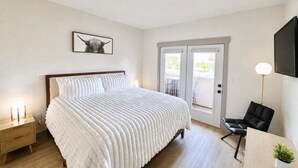 A hotel room with a bed, bedside tables, a desk, a chair, a TV, and a door leading to an outdoor area. - 2073 | Designer King Villa | Full Kitchen | Pool (Midway)