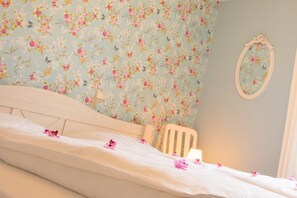 Una cama con cabecera de estampado floral, una silla blanca y un espejo montado en la pared.
