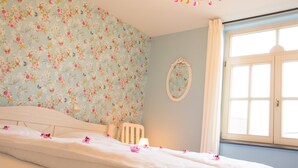 Ein Schlafzimmer mit floraler Tapete, einem Bett mit weißer Bettwäsche, einem weißen Sessel, einem Nachttisch mit Lampe und einem Fenster mit Vorhängen.