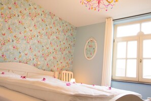 Een slaapkamer met bloemenbehang, een bed met witte lakens, een witte stoel, een nachtkastje met een lamp, en een raam met gordijnen.
