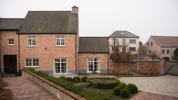 Ein Innenhof mit einem Backsteinpfad, einer rundlichen Hecke und einem Backsteinhaus mit zahlreichen Fenstern und einer flachen Dachfläche.