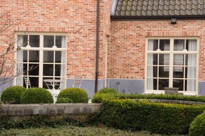 Een bakstenen gebouw met twee grote ramen, een kleine tuin met hagen en een ladder tegen de muur.