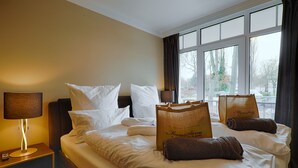 Habitación de hotel con cama, almohadas, lámpara de noche, ventana con cortinas y una maleta sobre la cama.