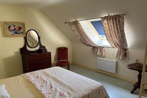A bedroom with a bed, a dresser, a mirror, a chair, a window with curtains, a radiator, and a small table with a plant. - Normandy Gite with Bay Views (Courtils)