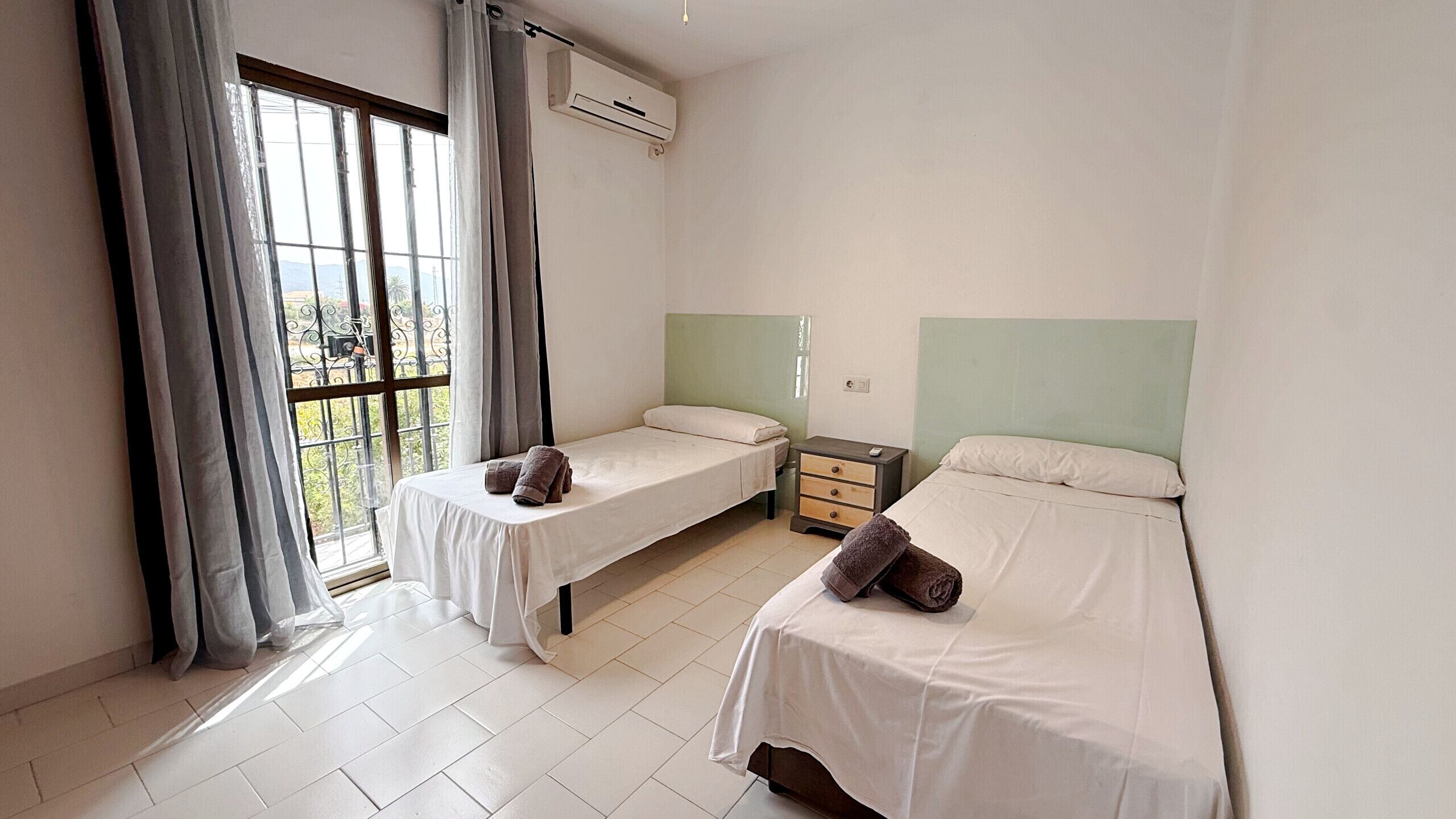 A room with two single beds, a small bedside table with a lamp, a window with curtains, and an air conditioning unit.