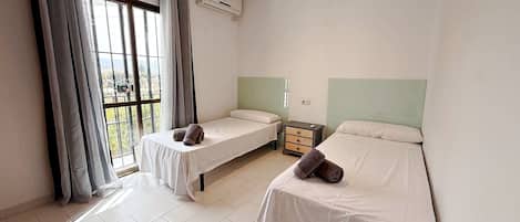 A room with two single beds, a small bedside table with a lamp, a window with curtains, and an air conditioning unit.