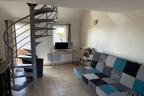 A living room with a spiral staircase, a sofa with patterned cushions, a television, and a small table with a vase of flowers. - Charming Normandy Escape for Three (Courtils)